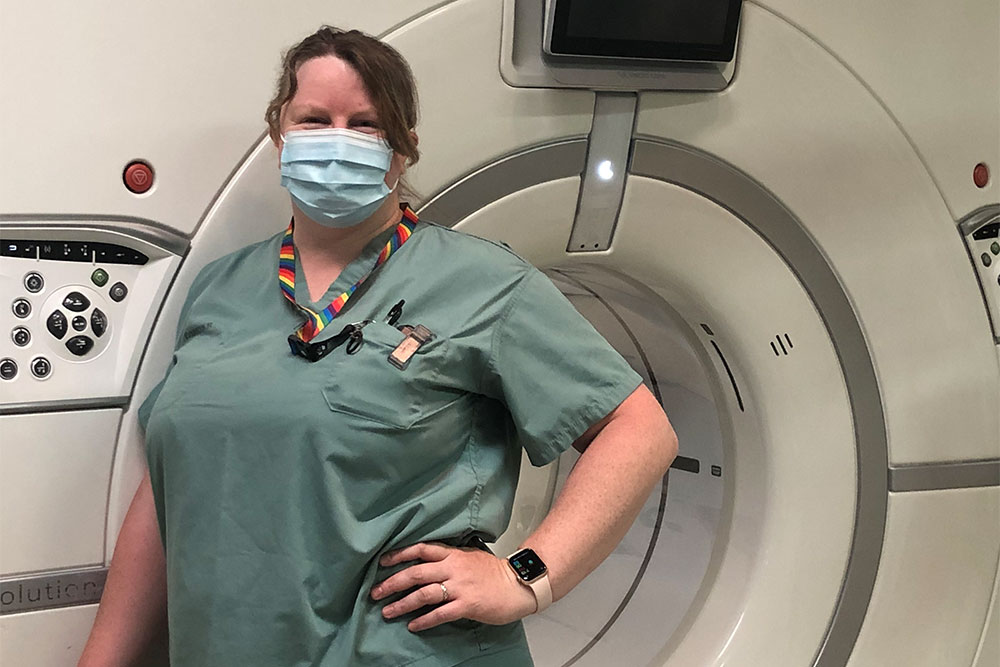 Helen Taylor, wearing scrubs and a mask, stands in front of a CT scanner.