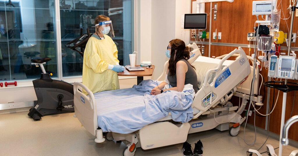 Emily gray sits up in a hospital bed and looks at Dr. Elizabeth Tullis, dressed in full personal protective equipment including a mask, face shield and gown.