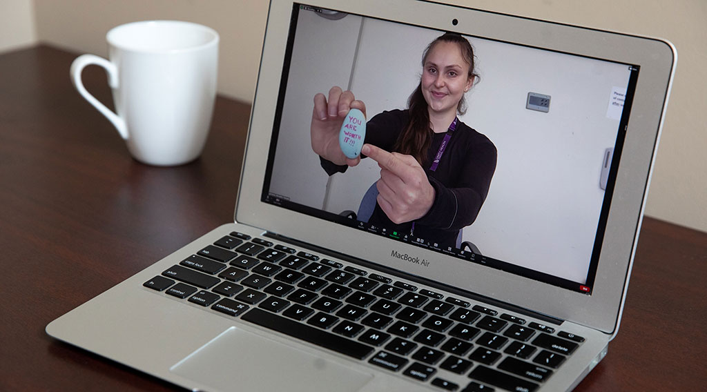 As seen on the screen of a laptop computer, Rachel Blair holds up a painted rock to a web camera during a Zoom call.