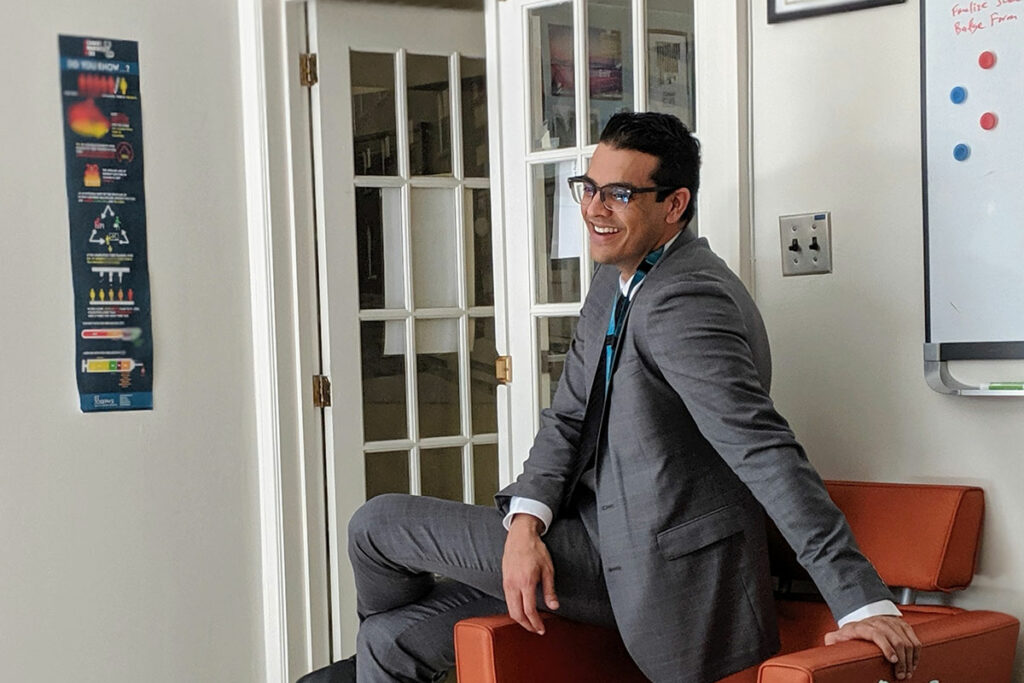 Jasdeep Grewal sits on the arm of a chair while speaking with someone outside the frame of the picture.