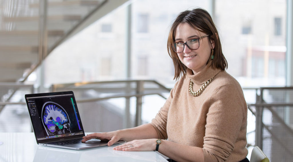 Dr. Katie Dunlop sits at a table in the common area outside the Health Sciences Library, looking at the camera and displaying a laptop computer showing an image scan of a human brain.