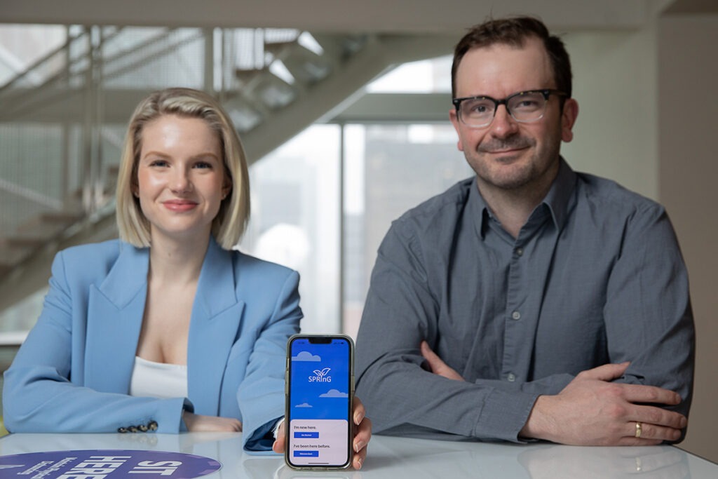 Madison Ford and Dr. Arthur McLuhan sit at a table in the Li Ka Shing Knowledge Institute. Ford is holding a smartphone with the SPRinG app displayed.