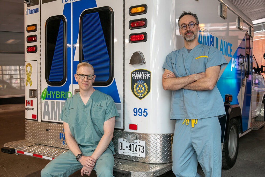Dr. Brodie Nolan sits on the bumper of an ambulance while Dr. Johannes von Vopelius-Feldt stands beside the ambulance to Dr. Nolan's left.